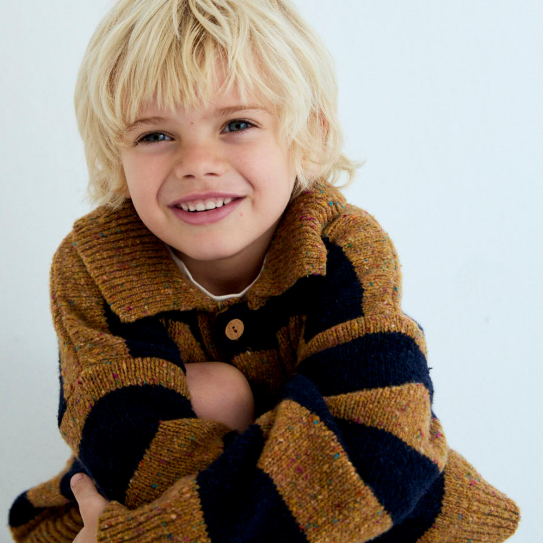 Sold at Bombon shop, this kids' knit polo sweater by The New Society features wide mustard and navy stripes, a classic collar with button placket, and a small embroidered chest logo—blending cozy comfort with a sporty, vintage-inspired look. model