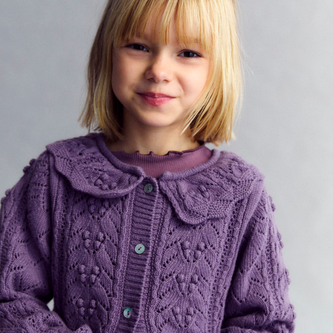Sold at Bombon shop, this girls’ cardigan by The New Society features a rich lilac hue with an intricate pointelle knit pattern, scalloped Peter Pan collar, and front button closure—offering a refined and feminine layer for cooler days. model