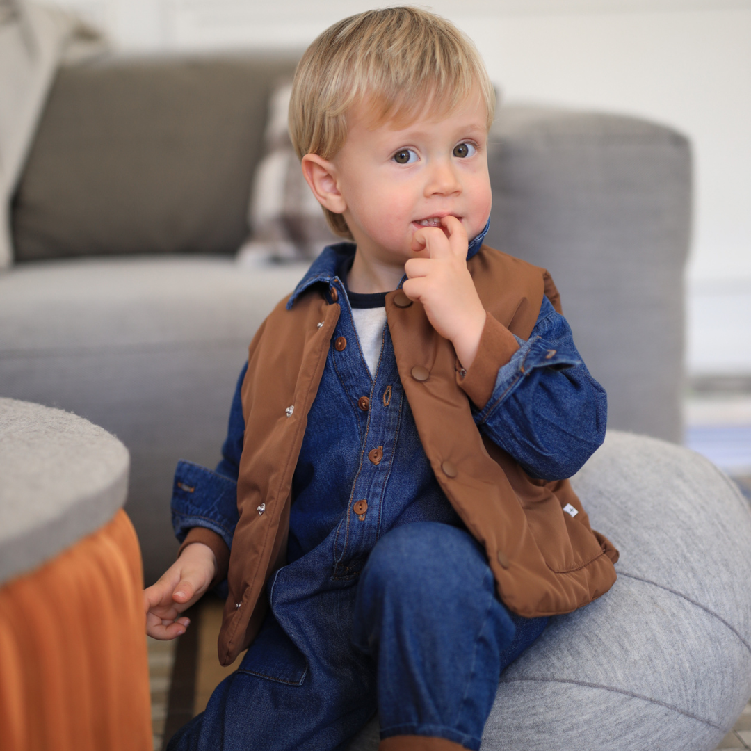 Sold at Bombon shop, designed by 1+ in the family — brown padded baby vest with front patch pockets and snap-button closure, laid flat on a white background. model