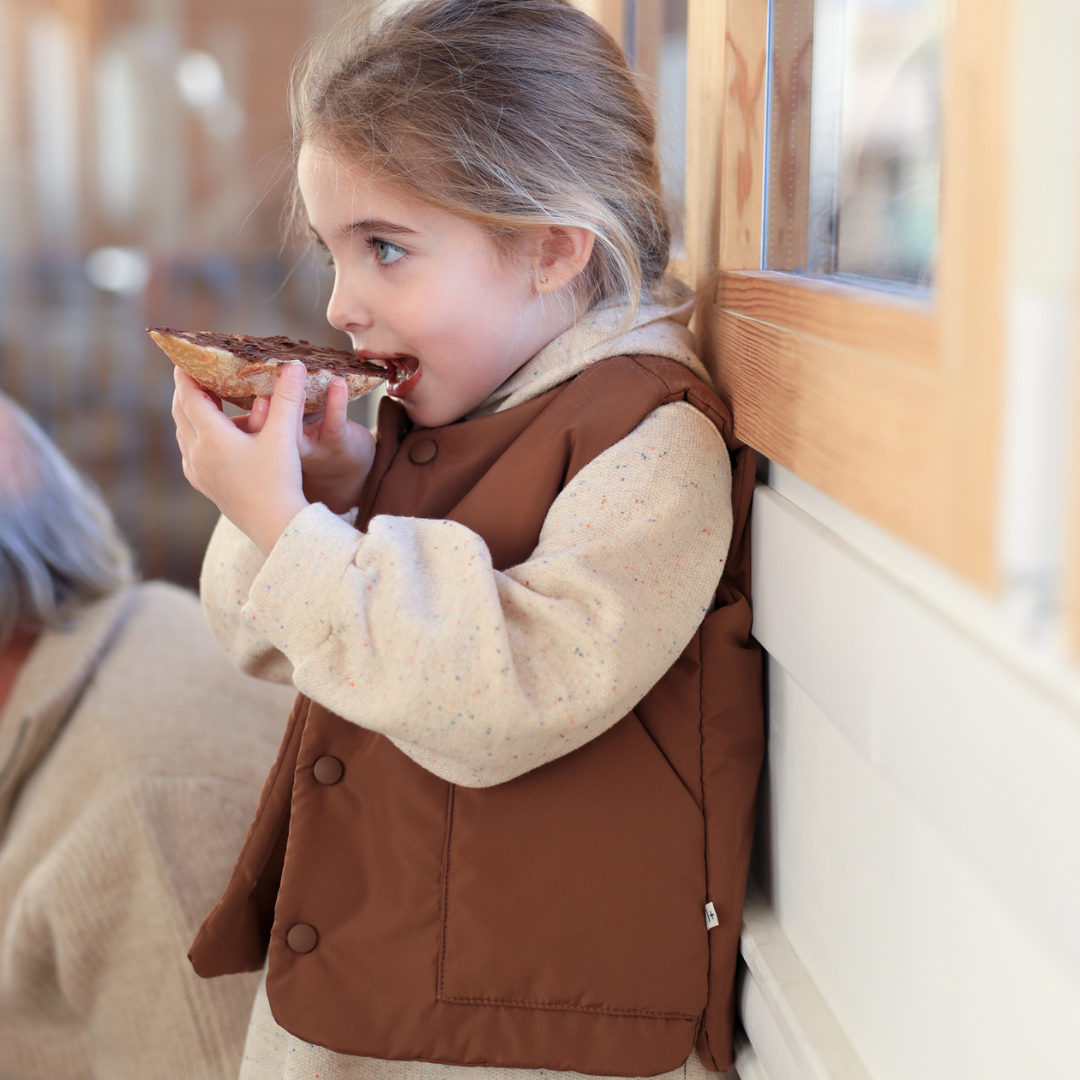Sold at Bombon shop, designed by 1+ in the family — brown padded baby vest with front patch pockets and snap-button closure, laid flat on a white background. model
