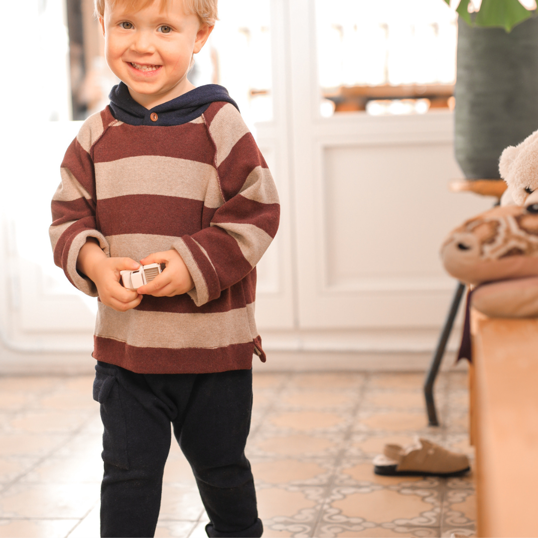 Child wearing a striped sweater standing indoors with a teddy bear in the background