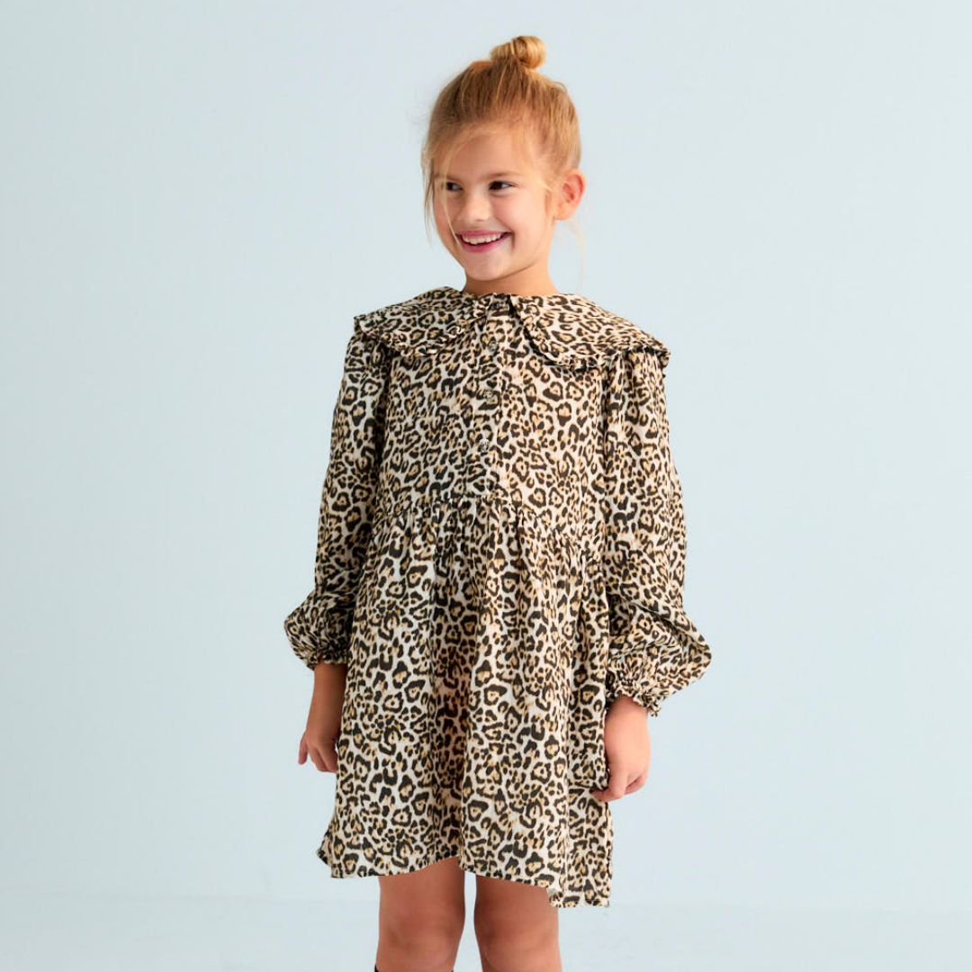 Sold at Bombon shop, this girls’ dress by The New Society features an all-over leopard print, ruffled collar, long sleeves with gathered cuffs, and a tiered silhouette for a bold and playful look. model front view