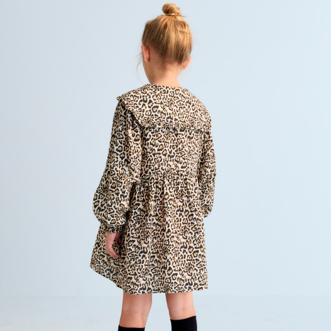 Sold at Bombon shop, this girls’ dress by The New Society features an all-over leopard print, ruffled collar, long sleeves with gathered cuffs, and a tiered silhouette for a bold and playful look. model back view