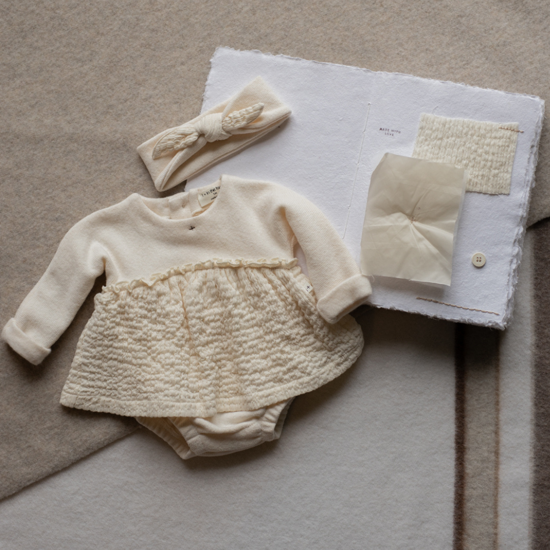 Sold at Bombon shop, designed by 1+ in the family — cream long-sleeve baby dress romper with ruffled gauze skirt and knit bodice. lifestyle