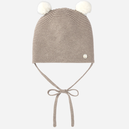 Sold at Bombon shop, designed by Paz Rodriguez, beige knit baby beanie with ivory pom-poms and chin ties.