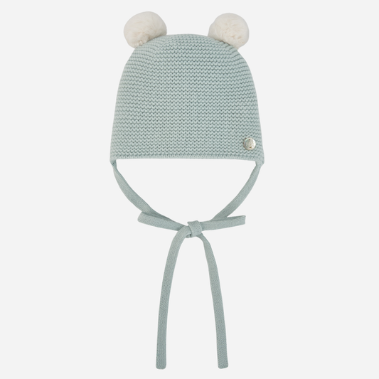 Sold at Bombon shop, designed by Paz Rodriguez, mint green baby knit beanie with two white pom-poms and chin ties.