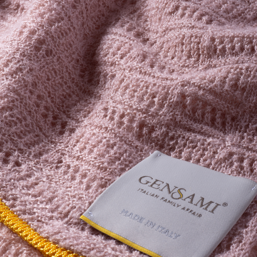 Sold at Bombon and designed by Gensami, a pink knitted baby blanket with openwork texture and a contrasting yellow trim along the border.